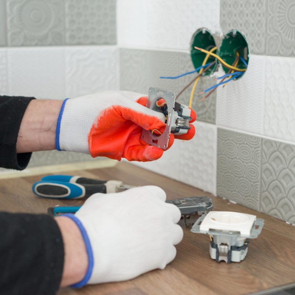 Renovation and construction in kitchen, close-up of electricians hand installing outlet on wall with ceramic tiles using professional tools.