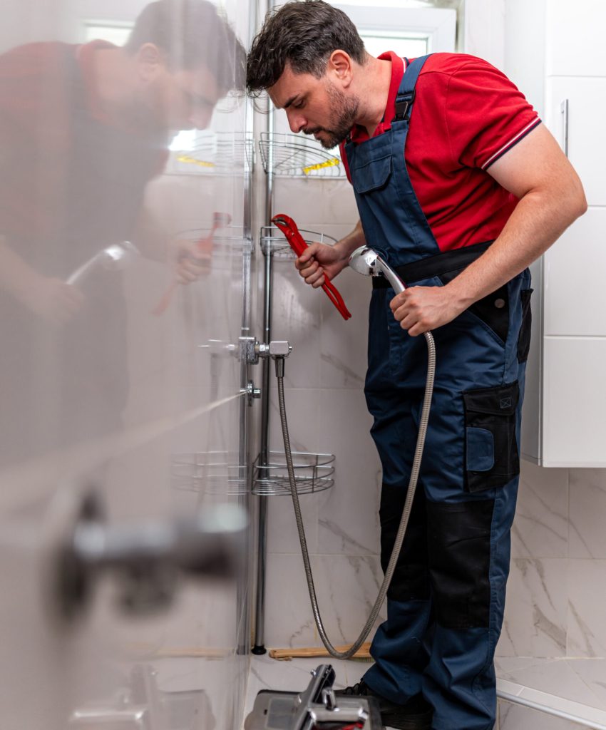 A plumber in a red shirt and blue overalls works on fixing a shower in a clean and bright bathroom.