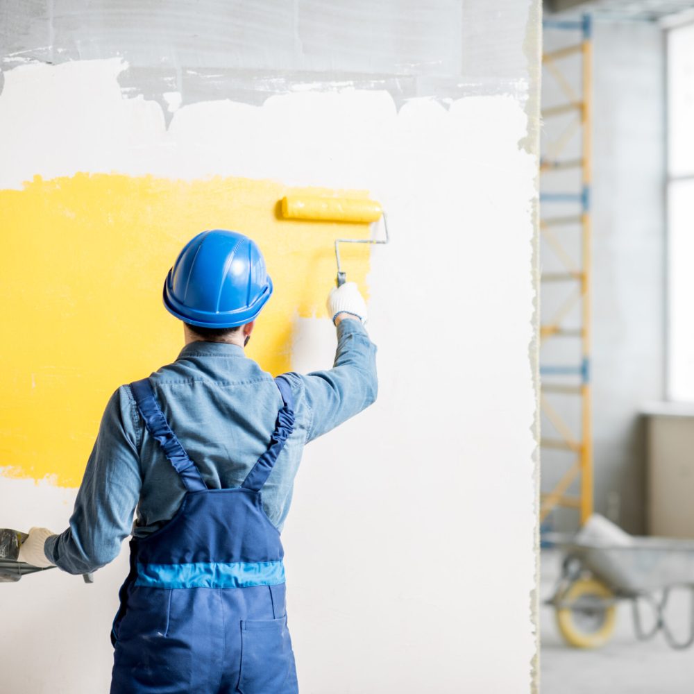Workman in uniform painting wall with yellow paint at the construction site indoors